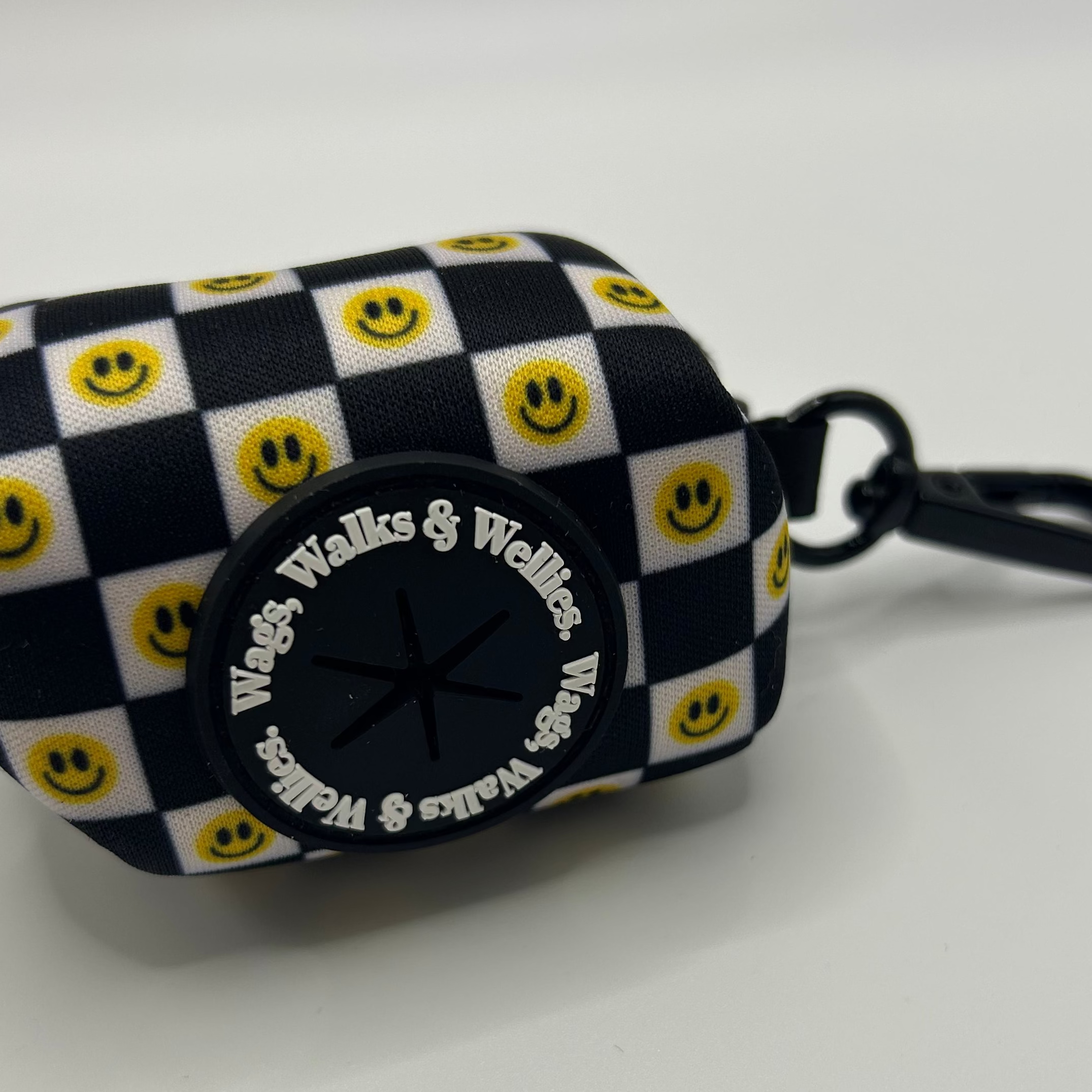 A small fabric pouch with a black and white checkered pattern and yellow smiley faces, featuring a black circular patch with the text 'Weds, Walks & Wednes' and a black key ring attached.
